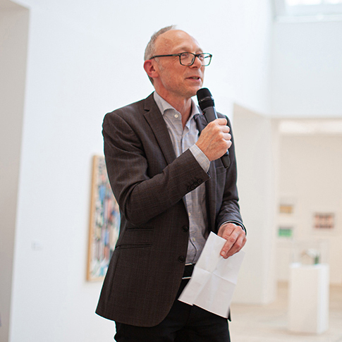 En halvkroppsbild på konsthallschef Mats Stjernstedt i kostym. Han bär glasögon och håller i en mikrofon i ena handen.