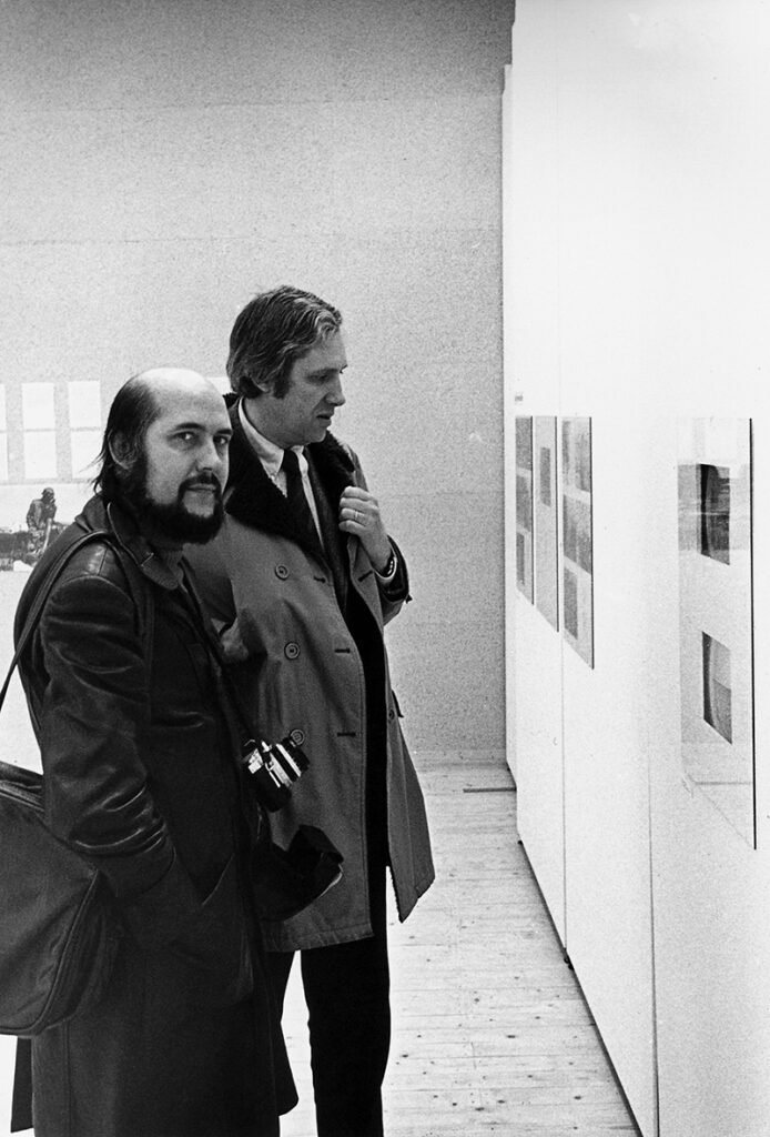 Svartvit bild av två män som står och tittar på inglasade verk på en vägg. En av männen kikar in i kameran.