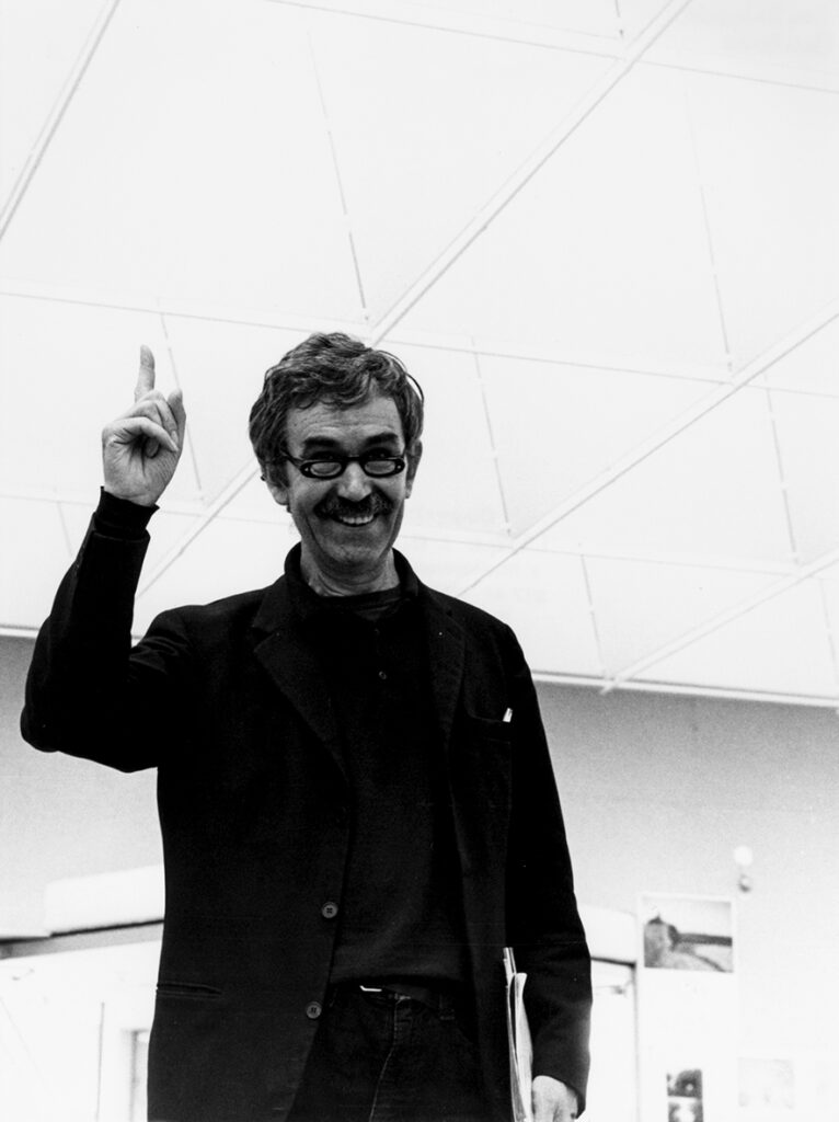 Ett halvkroppsporträtt av en man som ler, håller några papper i vänster hand och pekar upp i taket med höger hand.