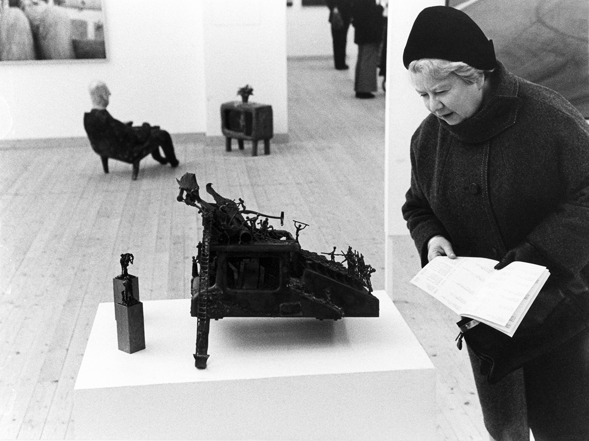 En äldre dam i mössa och tjock vinterkappa håller i en pamflett och tittar på ett konstverk