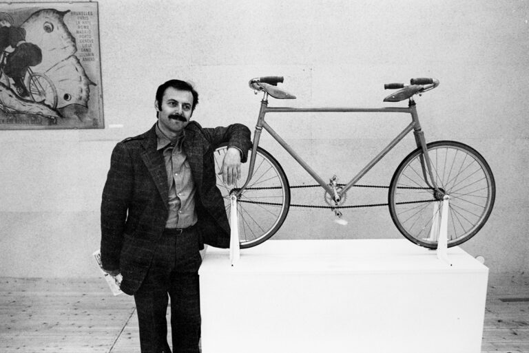 En man i kostym står lutad mot ett konstverk. Konstverket är en cykel med dubbel uppsättning sadel och styre samt ingen pakethållare.