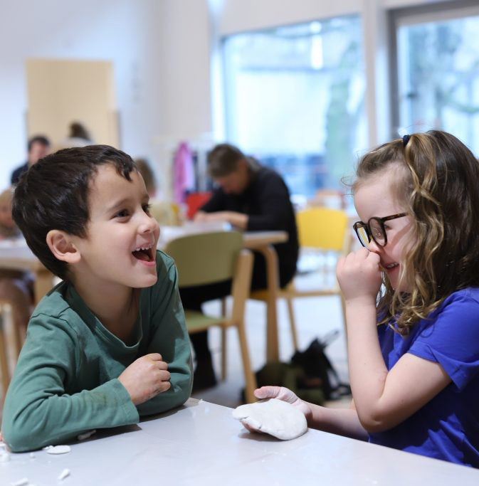 Två barn sitter vid ett bord, tittar mot varandra och skrattar. Ett av barnen håller en bit lera. I bakgrunden syns fler bord där det sitter personer samt stora ljusa fönster.
