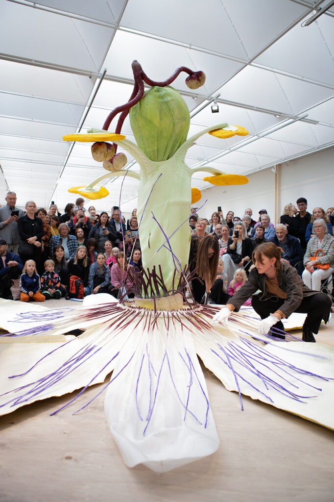 A large audience looks at a fantastic textile sculpture of a passion flower.