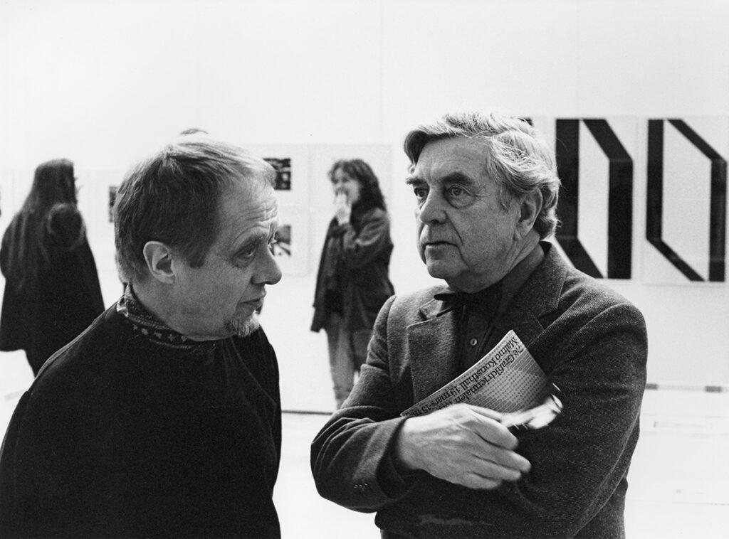 Två personer i samtal. En håller i en utställningskatalog.I bakgrunden syns två andra besökare samt några tavlor på väggen.