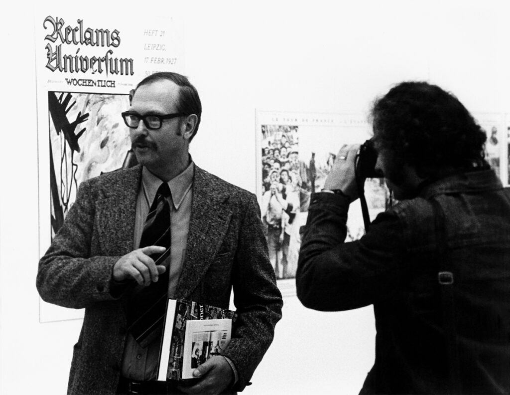 En person iklädd kostym, glasögon och hållandes några böcker blir fotograferad av en person till höger i bild.