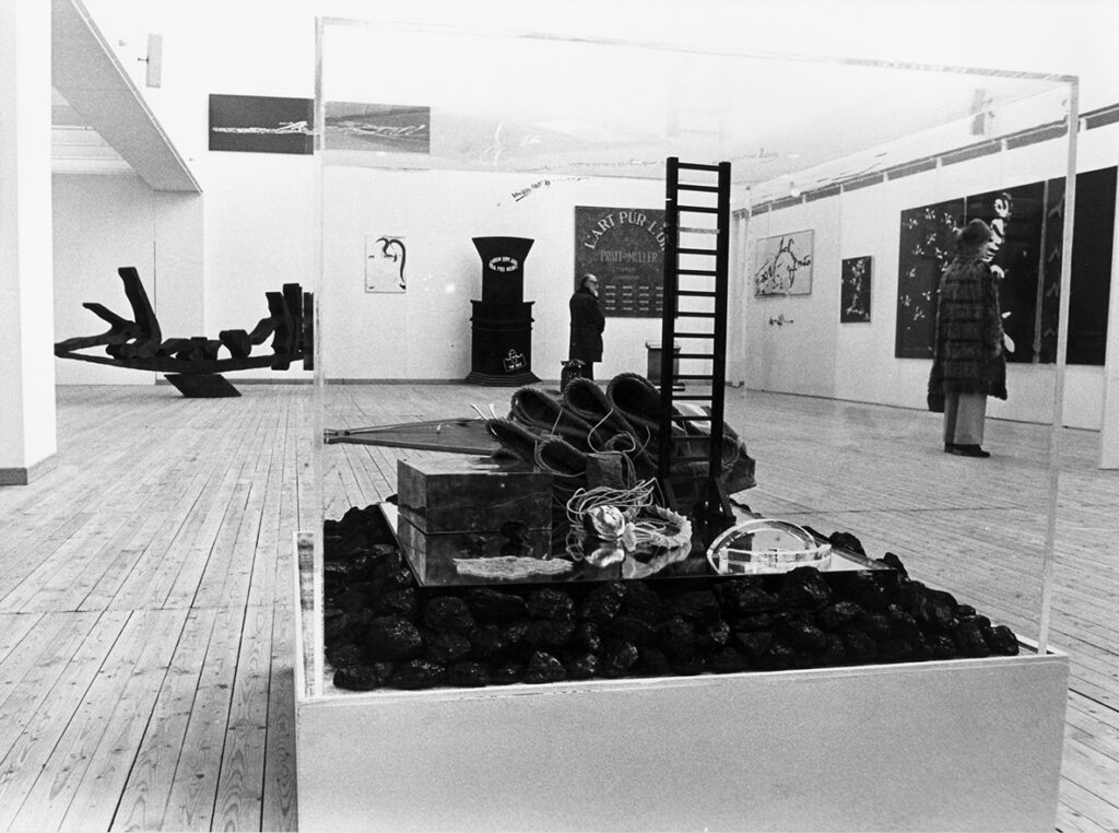 Utställningshallen under Carl Fredrik Reuterswärds utställning. Ett konstverk står utställt på golvet centrerat i bild. I bakgrunden syns konstverk både på väggar och golv samt två besökare.