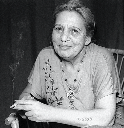 Svartvitt porträtt av Ceija Stojka som ler in i kameran. Tatuerade nummer syns på hennes arm.