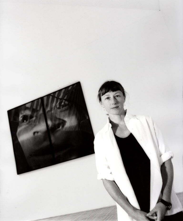 Cindy Sherman iförd svart tröja och vit skjorta framför ett fotografi på väggen. Fotograferad lite snett under ifrån.