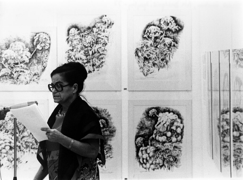 En person står vid en mikrofon och läser upp från ett papper. I bakgrunden syns sex större tavlor.