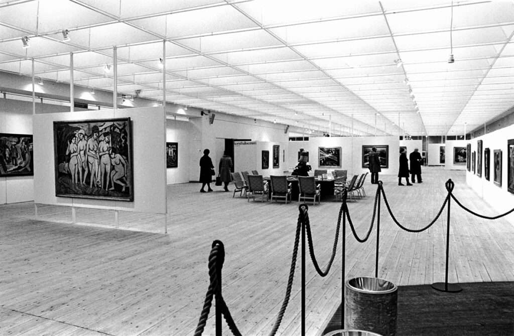 Konsthallens utställningshall med många målningar på väggarna. Några besökare kan ses lite längre bak i bild.