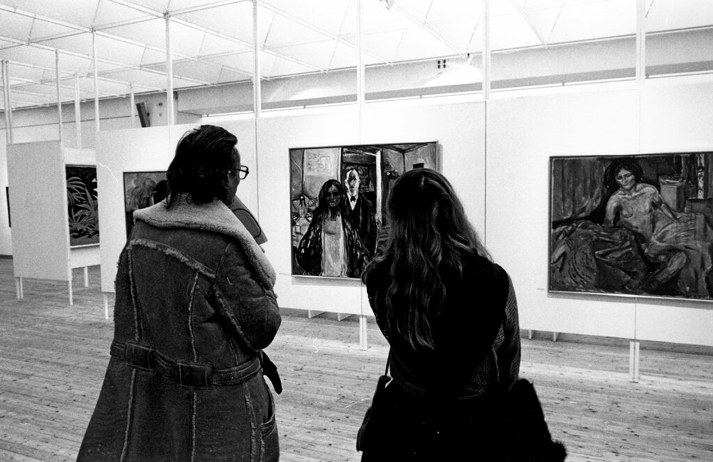 Två personer med ryggen mot kameran står och tittar på konstverk upphängda på en vägg.