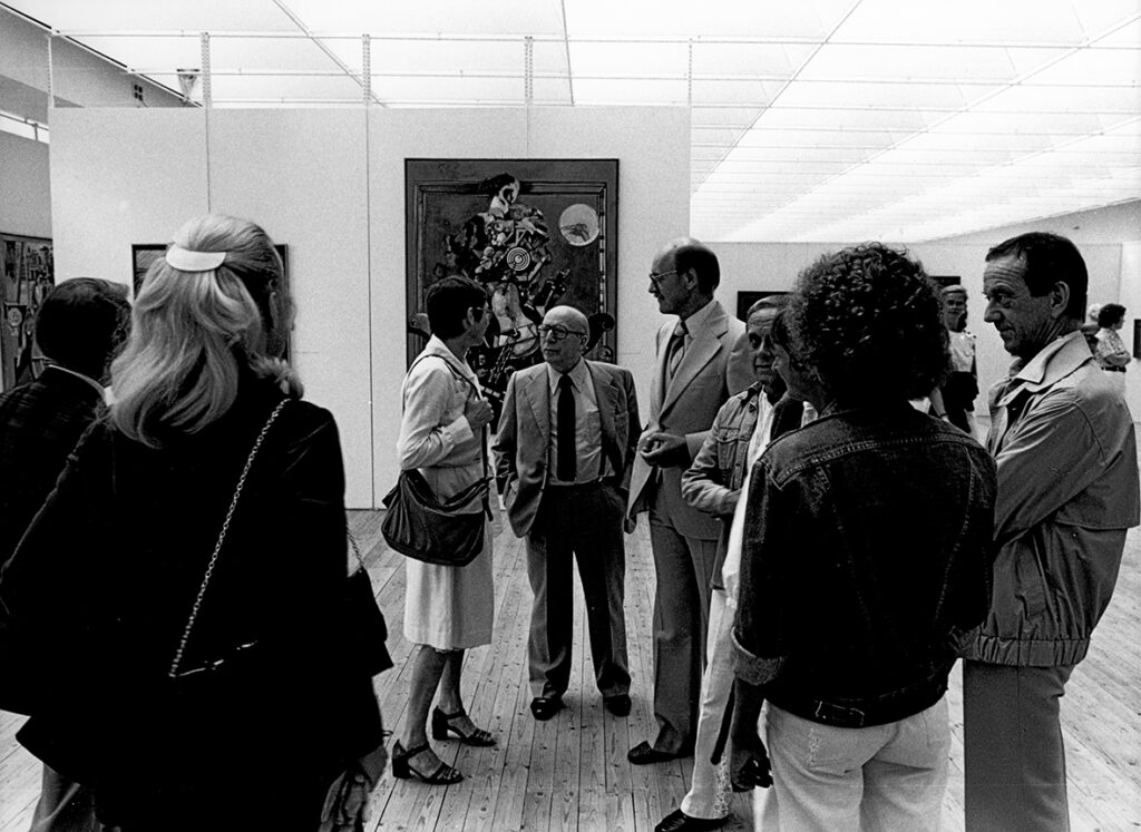 Svartvit bild på en samling människor i utställningshallen som står i ring. I bakgrunden syns tavlor på väggarna.