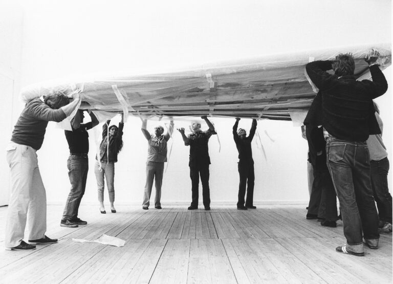 Tio personer står inne i utställningshallen och lyfter tillsammans upp en jättestor tavla som är inlindad i plast.