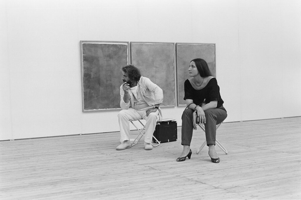 En man och en kvinna sitter på uppfällbara stolar inne i utställningshallen. Centrerade i bild och bakom de har de triptyk-målning.