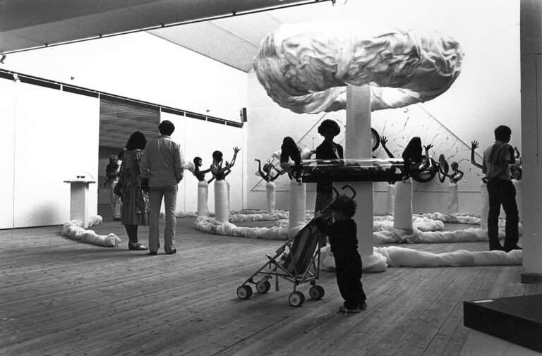 Bild från utställningshallen. Besökare kollar på ett stort konstverk. Konstverket består av ett stort moln i papper. Runt om står stora dockor.