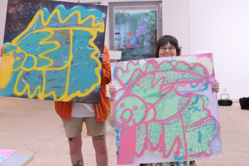 Two individuals holding up vibrant artworks. One person is holding the painting in a way that partially obscures them, showing only their legs, while the other is a joyful girl whose face peeks out from behind her artwork.