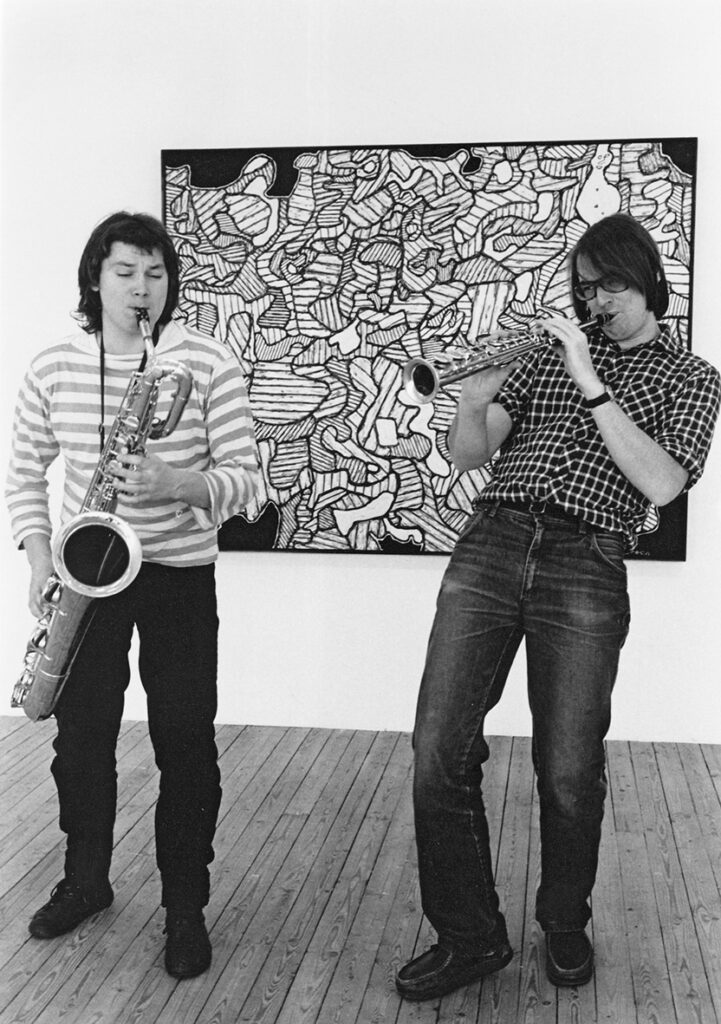 Två musikanter står framför en målning och spelar på sina blåsinstrument.