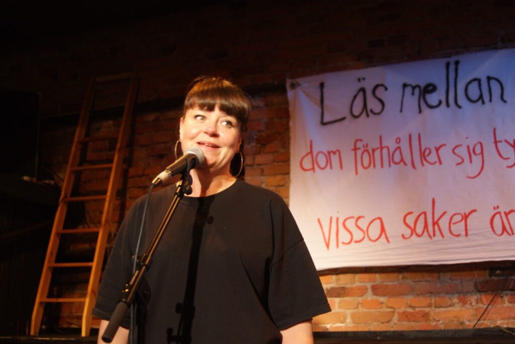 Konstnären Johanna Gustavsson framför ett plakat på en scen. Hon ler och talar i mikrofon.