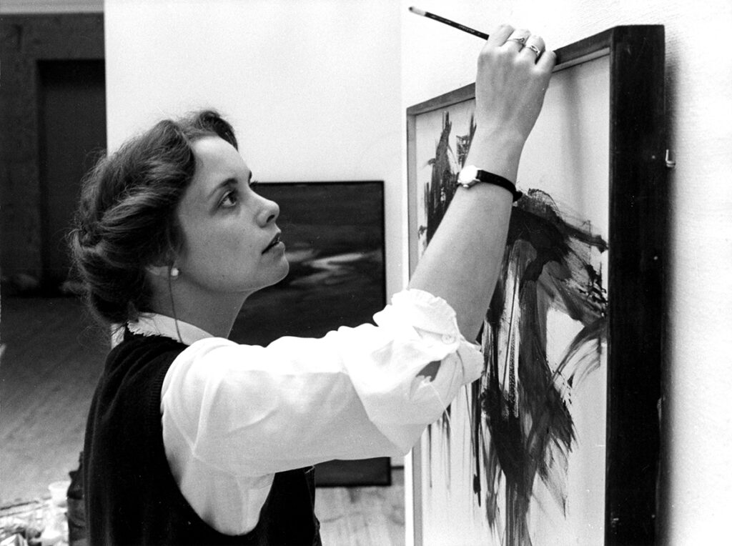 En kvinna står med en pensel i handen och tittar koncentrerat på en tavla.