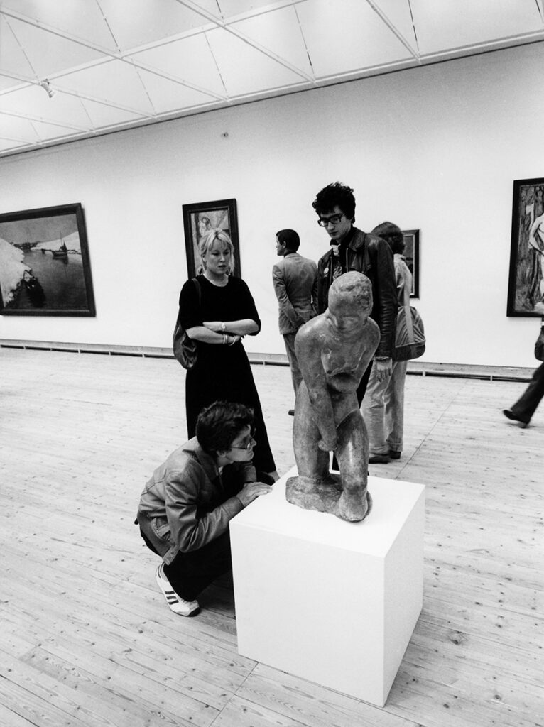 En skulptur står utställd i utställningshallen. Tre besökare står nära och undersöker den. I bakgrunden syns andra besökare och konstverk på väggarna.