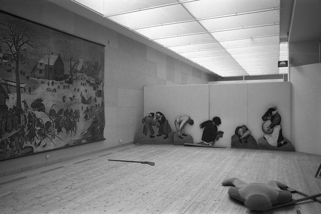 En svartvit bild från utställningshallen. På väggen är ett stort konstverk upphängt. Längs en annan vägg är figurklippta figurer uppsatta. På golvet ligger en kudde som ser ut att ha en människoform