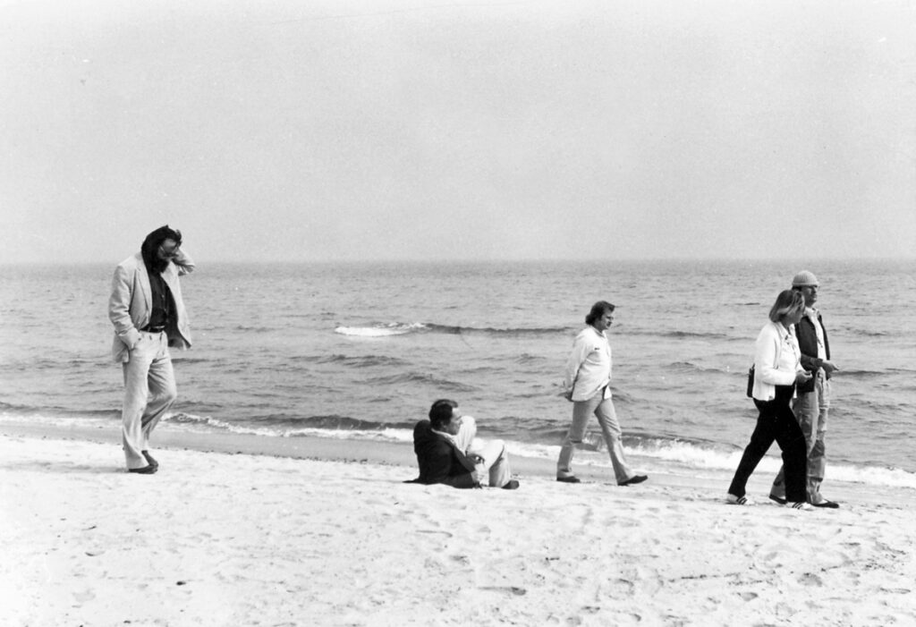 I bakgrunden syns havet och i förgrunden, på stranden, finns fem personer. Fyra personer står upp och går. En person sitter i sanden.