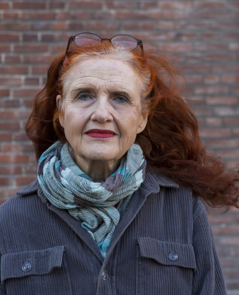 Konstnären Marianne Andersson Embäck framför en tegelvägg. Hon har rött hår, rött läppstift, glasögon och en sjal runt halsen. 