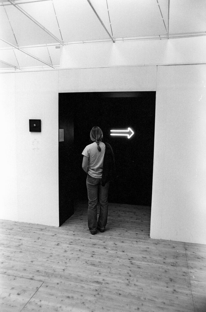 En bild från utställningshallen. En person står och tittar in i ett svart rum, med en lysande pil åt höger.