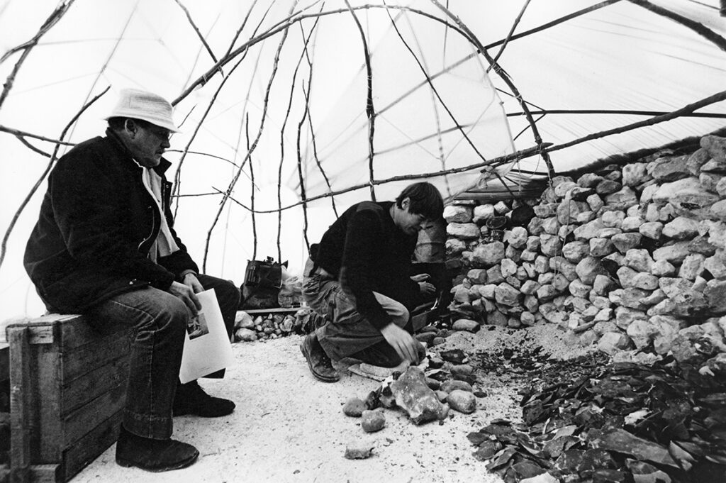 Två personer sitter i en tält-formation. En person sitter på en låda och tittar på den andra som staplar sten.