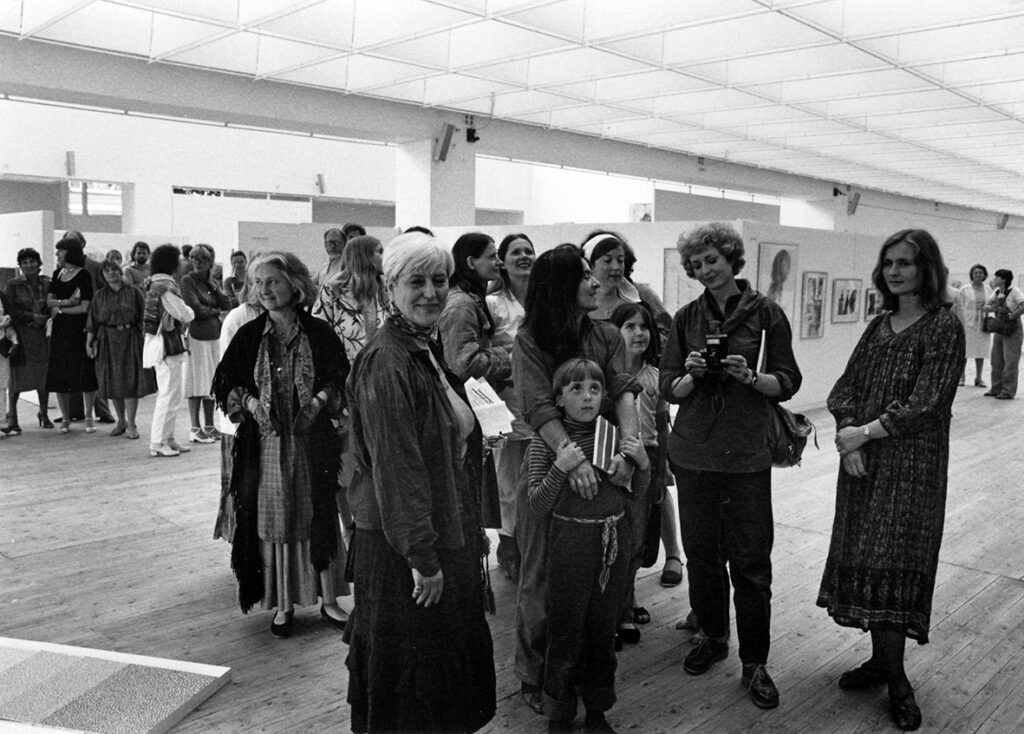 Bild från utställningshallen. En större grupp besökare står centrerat i bild och tittar mot kameran.
