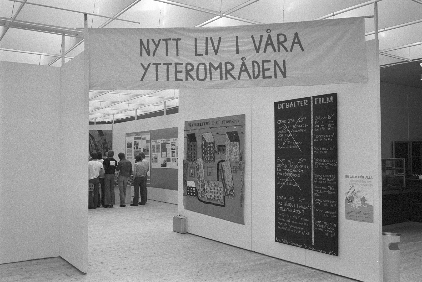 Picture from the exhibition space. A banner can be seen that reads "New life in our outlying areas". A number of visitors can be seen standing close to each other looking at something out of sight.