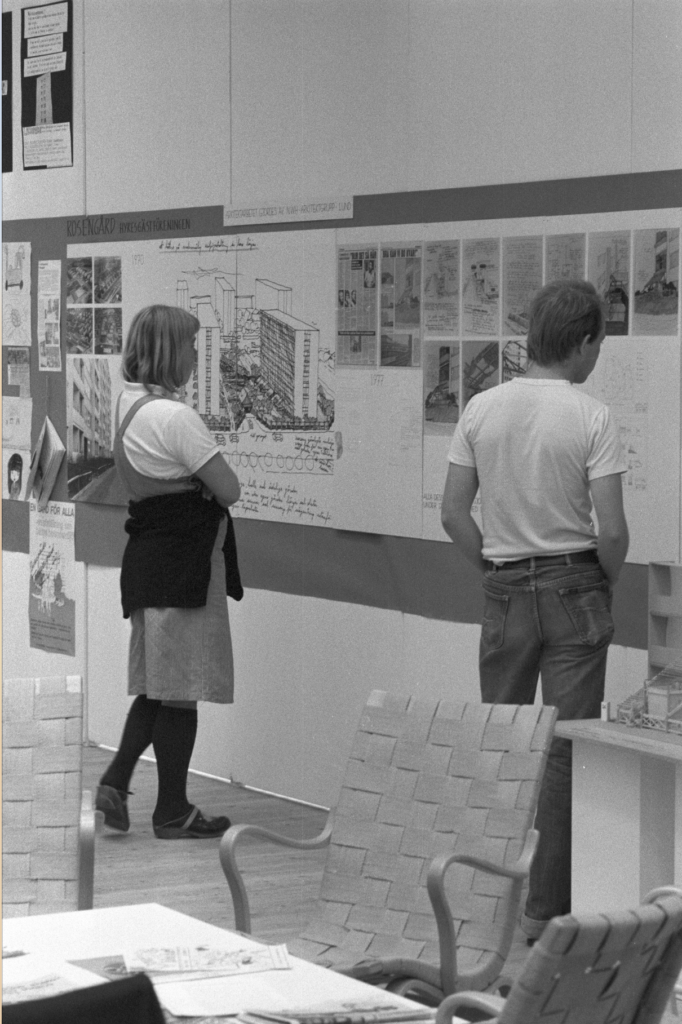 Två personer står och tittar på verk som är uppsatta på väggen. På väggen ses en illustration av hus samt diverse bilder. I förgrunden syns några fotöljer och ett bord.