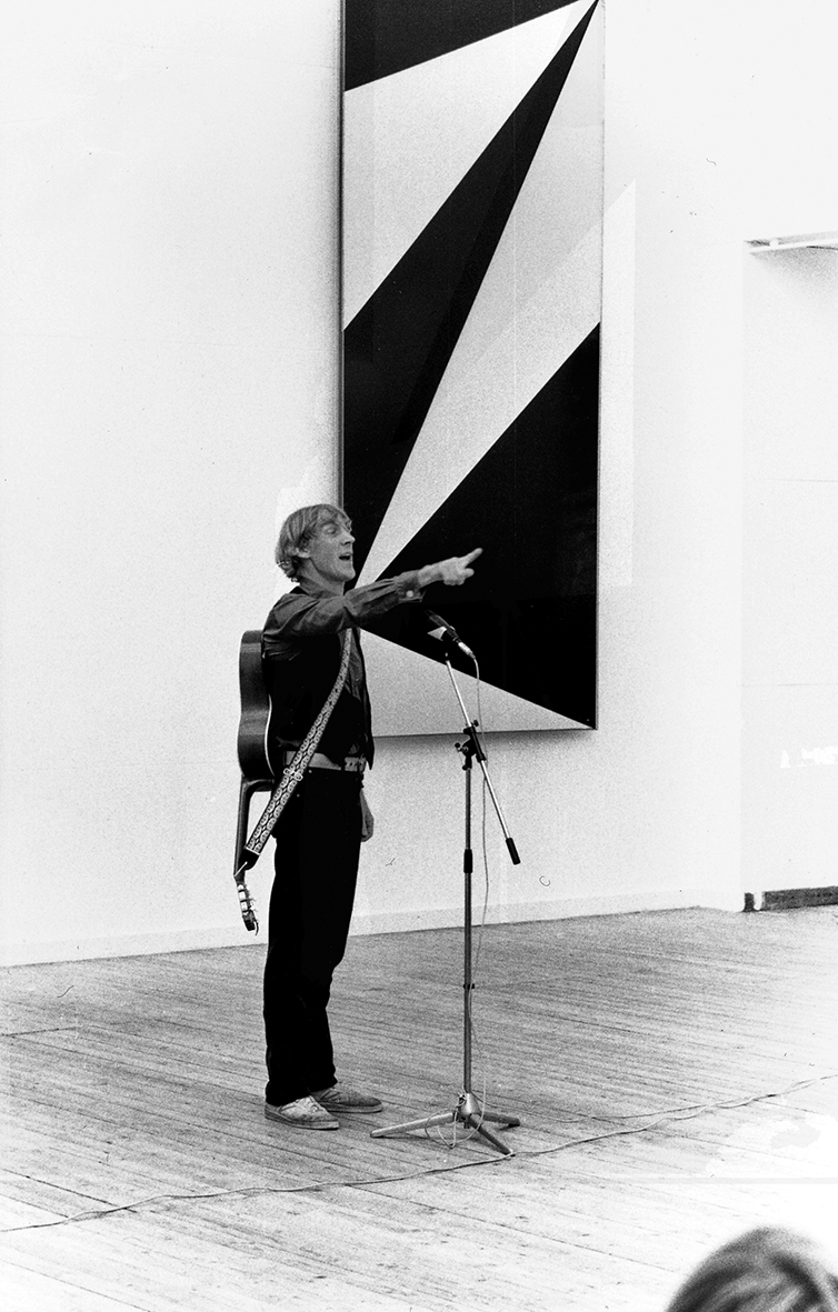 En person står vid en mikrofon med en gitarr på ryggen. Bakom honom hänger en tavla på väggen med geometriska former.
