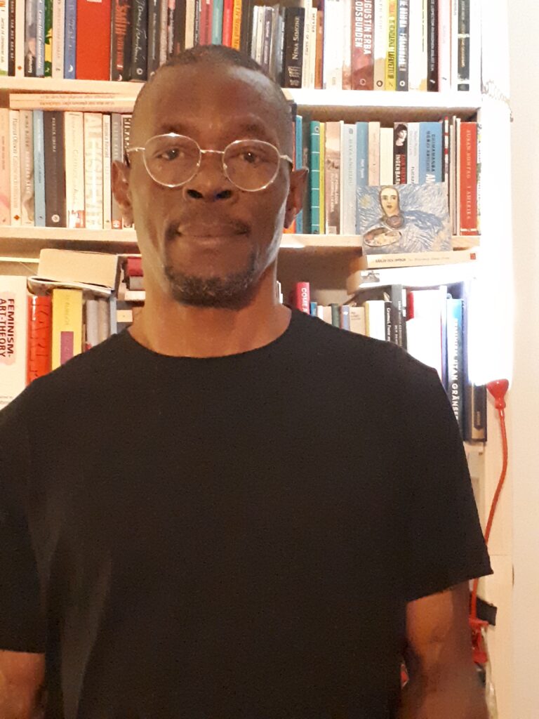 Konstnären Rex Okoro Ijomanta framför en bokhylla. Han är iklädd svarta t-shirt och metallbågade glasögon.