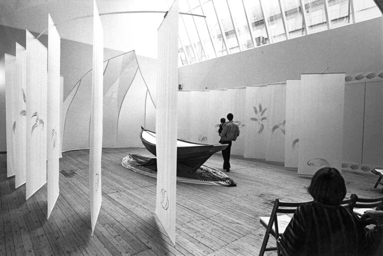 En båt står utställd mitt på golvet i utställningshallen. Runt om sitter det stora vita papper upphängda i en form runt båten. Tre besökare syns i bild.