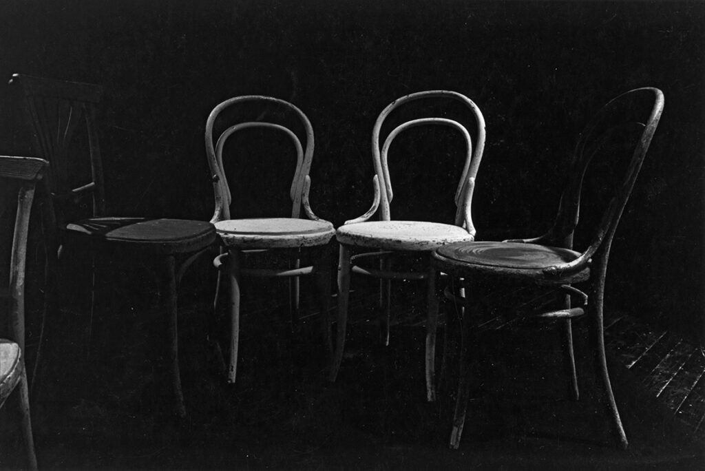 En svartvit bild på fyra trästolar som står på rad. De två i mitten är vitmålade och de på kanterna är i en mörkare ton. I vänster kant av bilden syns en bit av en annan stol.