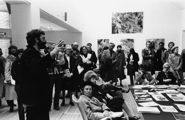 En stor grupp människor är samlade runt en man som talar i konsthallen. På väggarna hänger stora målningar.