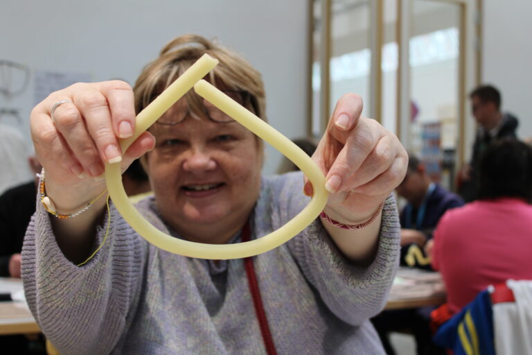 A woman in a knitted sweater holds up a yellow form of beeswax towards the camera with both hands. In the background, several other people can be seen creating.