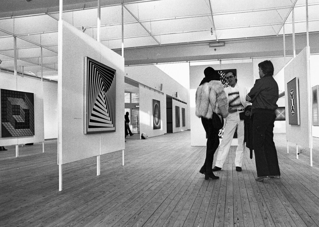 Tre besökare står och pratar med varandra i utställningshallen med tavlor på väggarna runtomrking.