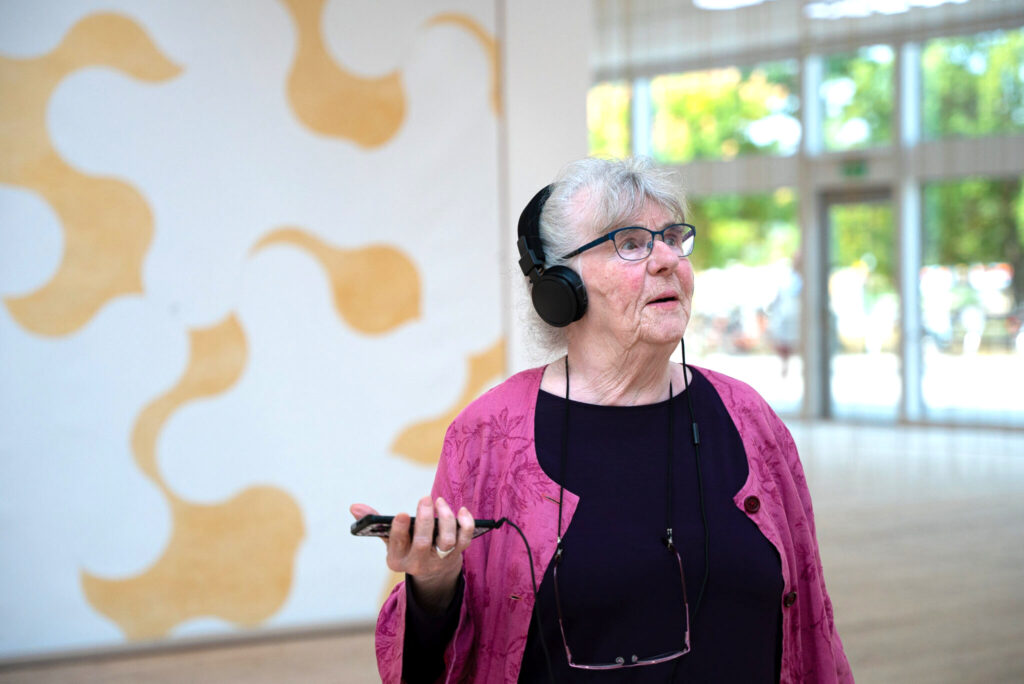 Äldre kvinna lyssnar på auidiguiden i en av konsthallens utställningar.