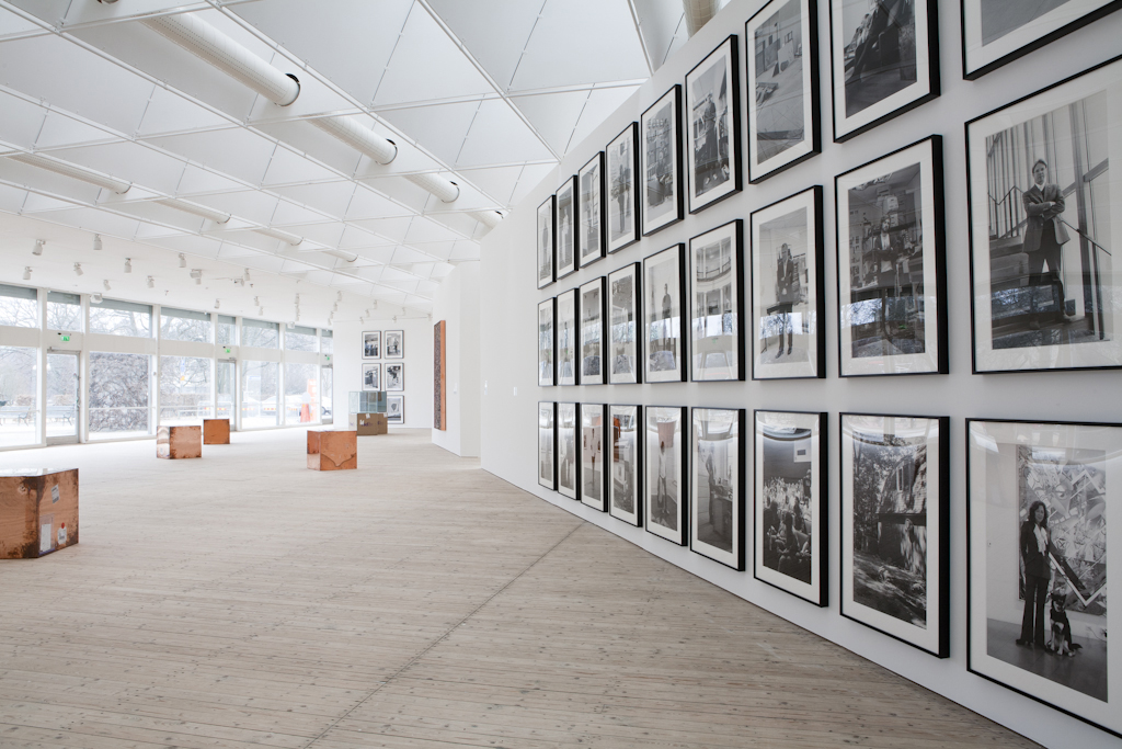En vägg med inramade fotografier löper in i konsthallen. Kopparglänsande kuber står utspridda på golvet.