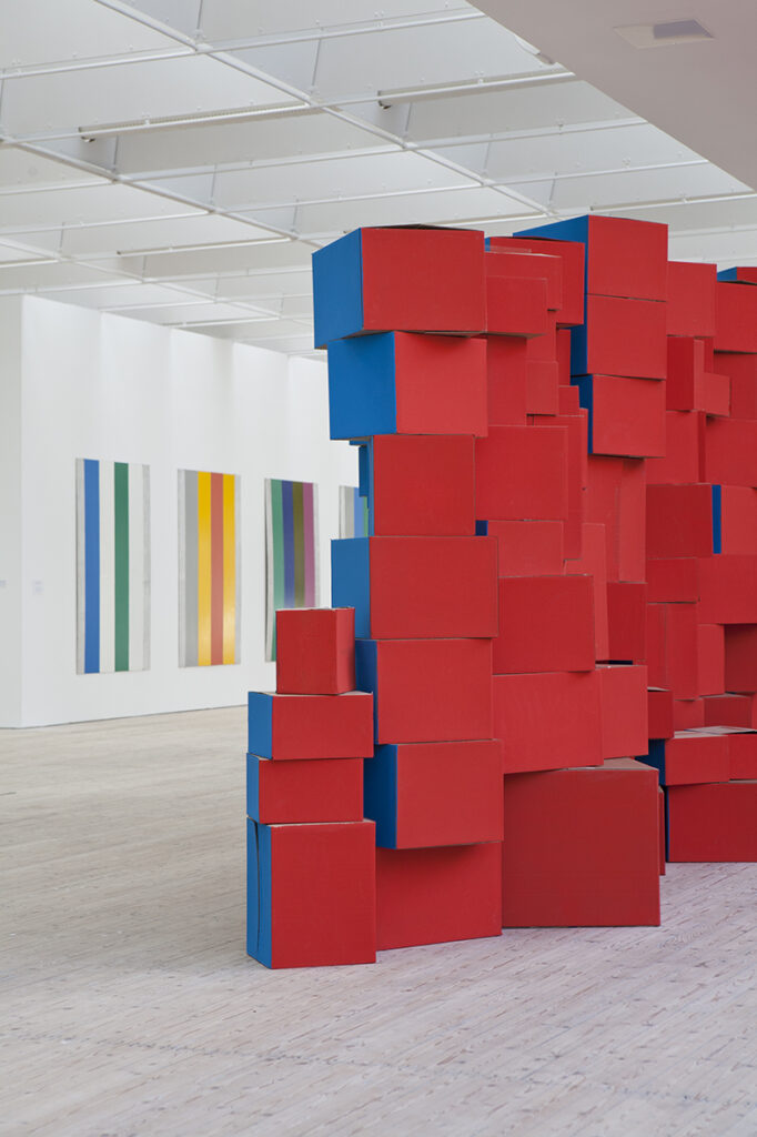 Bild från konsthallsrummet med en geometrisk installation i starkt rött och blått. I bakgrunden skymtar grafiska verk på väggen.