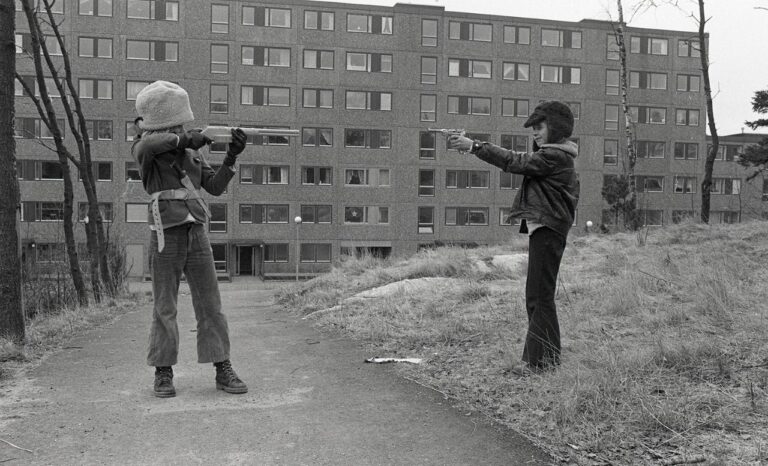 Två personer siktar mot varandra med gevär framför ett höghus, bilden är svartvit.