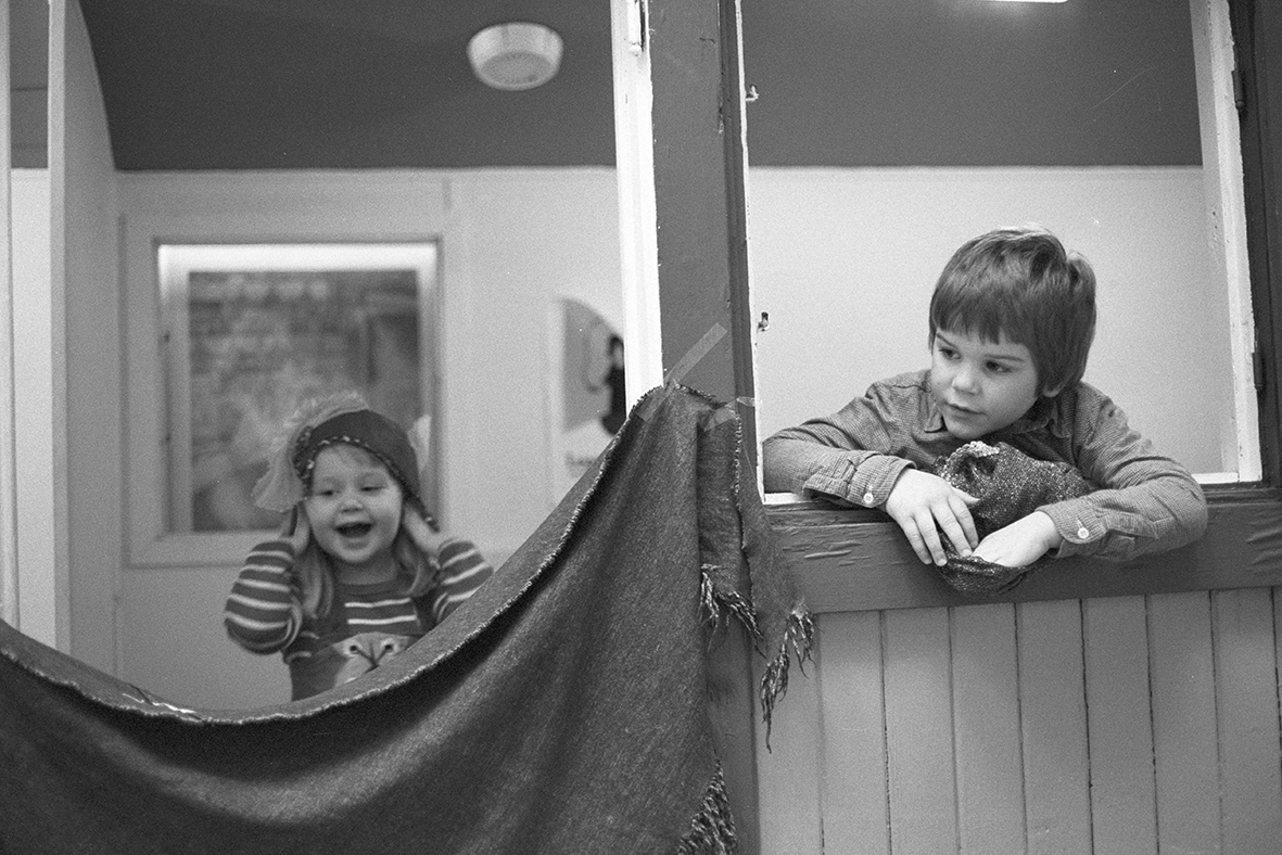 Details of a playhouse. A child to the right is looking out from a window. On the left side of the image, another child is a bit further back behind a hung-up sheet. She looks happy.