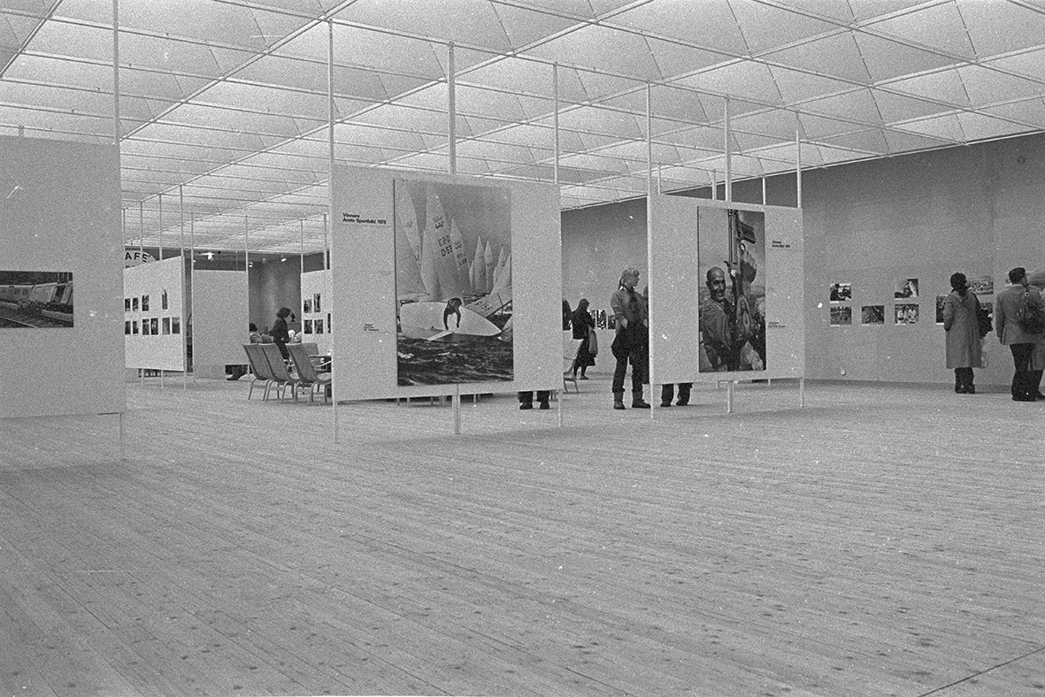 An image from inside the exhibition hall. Ceiling-mounted boards with pegs are in focus, displaying large photographs. In the background, visitors can be seen looking at art.