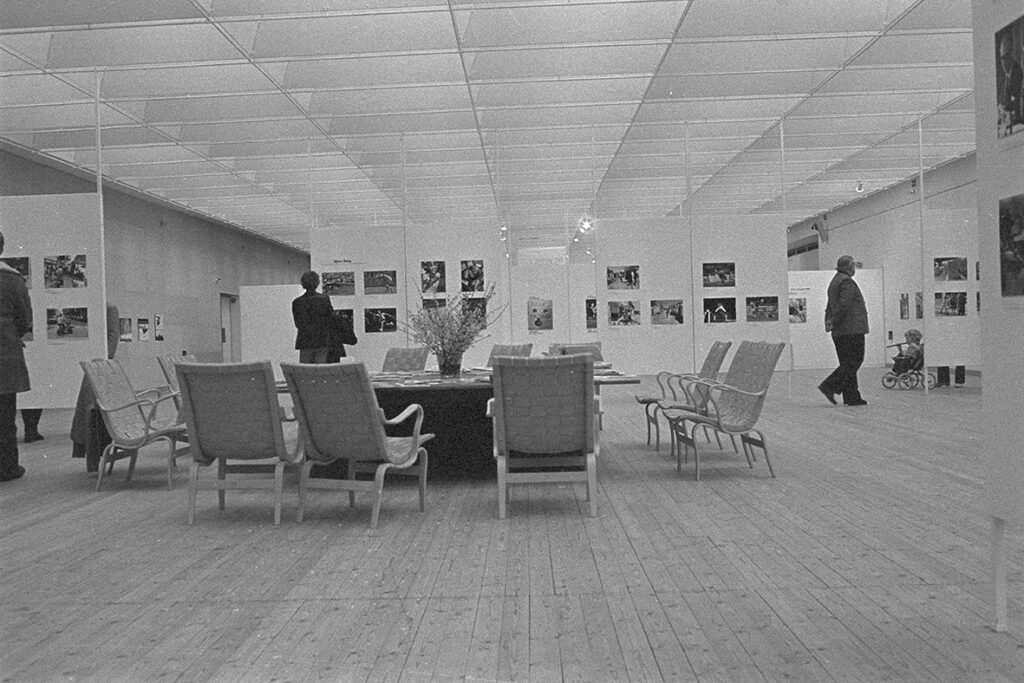 Ett stort bord och ett tiotal fåtöljer står i mitten av rummet. I bakgrunden ses konst på väggarna och några besökare beundra denna.