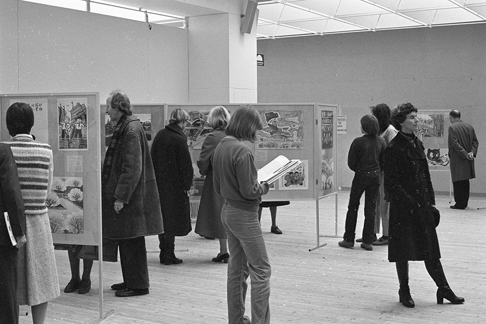 Image from inside the exhibition hall. Around ten visitors are standing and looking at larger wooden boards on the ground. Drawings and texts are displayed on these boards.