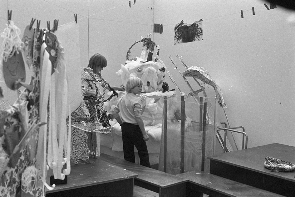 Two children stand around a large sculpture. In the upper part of the image, there are strings with wooden clips, and a child's drawing is hanging on them. In the foreground, tables and more children's drawings and other art are attached with clips.