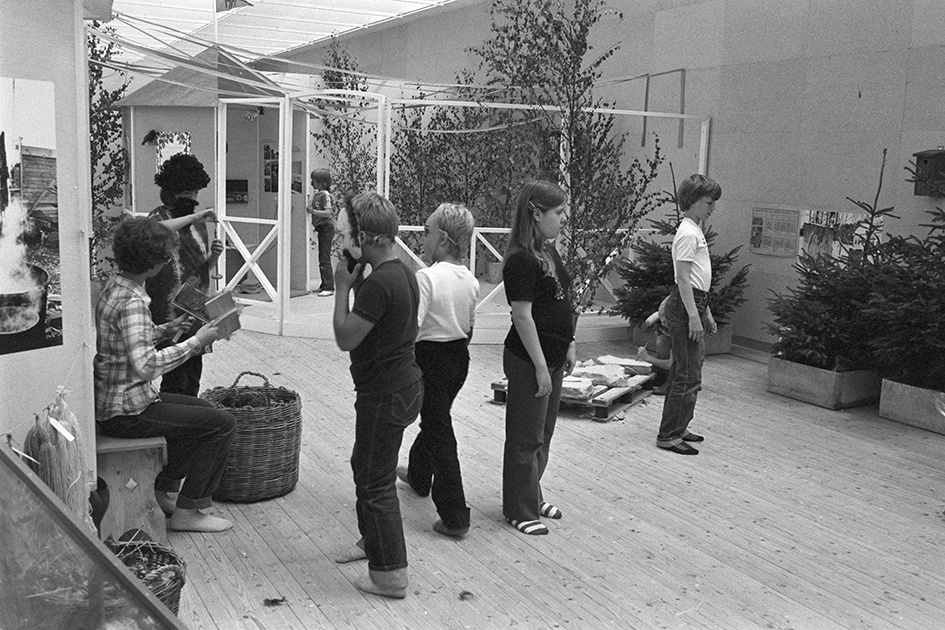 Bild inifrån utställningshallen. Centrerade i bild syns sex stycken barn stå och leka, några iförda utklädningskläder. I bakgrunden syns ett staket och några träd.