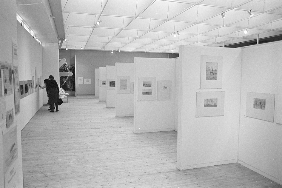 Bild inifrån utställningshallen. Tavlor och mindre bilder uppsatta längs väggarna. Två personer en bit från kameran står och tittar mot ett konstverk.